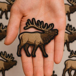 May include: An embroidered patch featuring a brown elk with large antlers, depicted in a side profile view. The elk has a tan body, dark brown neck fur, and a black stitched border outlining the entire shape of the animal against a light background.