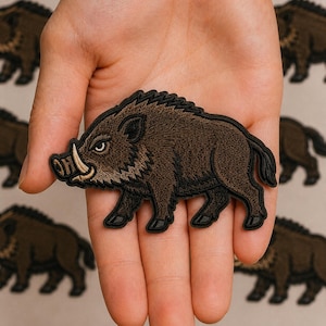 May include: An embroidered patch featuring a brown wild boar in profile, depicted with dark fur, a prominent curved tusk, and a black border. The design shows the animal standing on all four legs, with detailed stitching creating texture and depth.