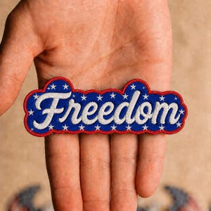 May include: An embroidered patch featuring the word Freedom in white cursive script on a blue background. The patch has a scalloped red border and is decorated with small white stars scattered across the blue field, creating a patriotic design.