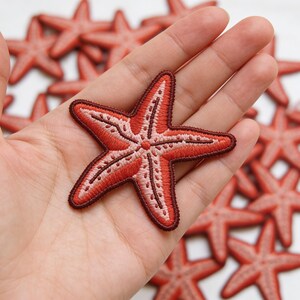 May include: Embroidered red starfish patch with a detailed design. The patch features a lighter red center and a darker red outline. Several similar patches are visible in the background.