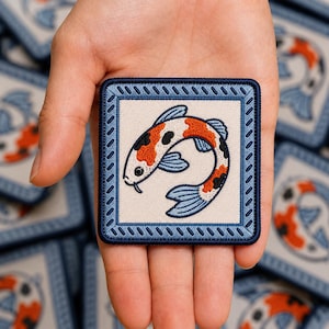 May include: A square embroidered patch featuring a white background with a blue border and a central koi fish design. The koi fish is depicted in orange, black, and white with blue fins, curved in a circular motion within the square frame.