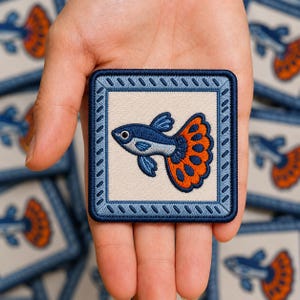 May include: A square embroidered patch featuring a guppy fish with a blue body and a vibrant orange tail with dark blue spots. The fish is centered on a cream-colored background, framed by a decorative light blue border with a dark blue edge.