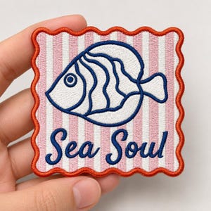 May include: A square embroidered patch with a scalloped orange border featuring a white and pink vertical striped background. The patch displays a white fish with dark blue outlines and the text Sea Soul in dark blue cursive script.