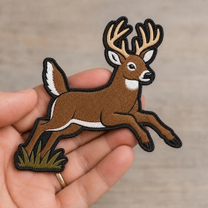 May include: An embroidered patch featuring a brown deer with tan antlers and a white tail, leaping over a patch of green grass. The design is outlined in black thread, showcasing detailed stitching of the animal's fur, facial features, and hooves.