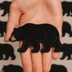 May include: An embroidered patch in the shape of a black bear walking. The bear is primarily black with a tan-colored snout and detailed stitching throughout the body, showcasing the silhouette of the animal in a side profile view.