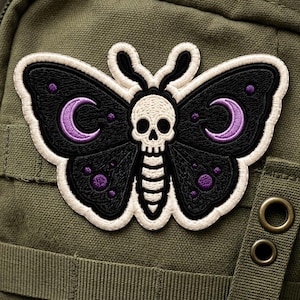 May include: An embroidered patch featuring a black moth with a white skull at its center. The wings are decorated with purple crescent moons and small purple dots, all outlined in a thick white border against a dark olive green fabric background.