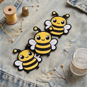 May include: Three embroidered bee patches featuring yellow and black striped bodies, white wings, and antennae. The patches are arranged on a blue denim fabric surface alongside a wooden spool of yellow thread, a metal thimble, and fabric glue.