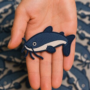 May include: An embroidered patch shaped like a catfish, featuring a dark navy blue upper body and a light cream-colored underbelly. The fish design includes detailed stitching, a visible eye, and long whiskers extending from the mouth area.
