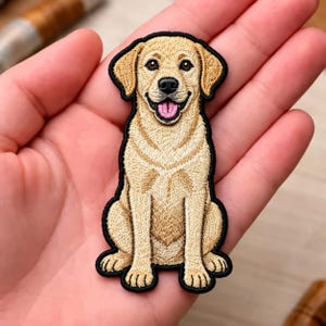 May include: An embroidered patch featuring a yellow Labrador retriever sitting in a forward-facing position. The dog has light tan fur, a black nose, and an open mouth with a visible pink tongue. The entire design is outlined in a thick black border.