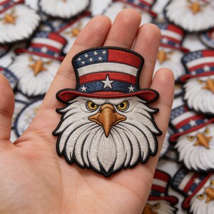 May include: Embroidered patch featuring an American bald eagle wearing a patriotic top hat. The hat is red, white, and blue with stars. The eagle has a stern expression and is surrounded by other patches.