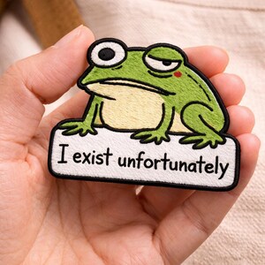 May include: Embroidered patch featuring a green frog with a grumpy expression, holding a white sign with the text "I exist unfortunately" in black. The frog has black eyes and a red cheek. The patch is likely for clothing or accessories.