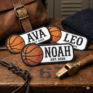 May include: Rectangular white patches with black text and a basketball design. The patches have the names AVA, LEO, and NOAH, with "EST. 2026" below each name. A leather strap and a brass whistle are also visible.