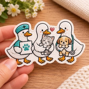 May include: An embroidered patch featuring three white ducks. The left duck wears a teal medical cap and shirt, holding a chart with a teal paw print. The middle duck holds a gray tabby kitten. The right duck wears a stethoscope and holds a puppy.