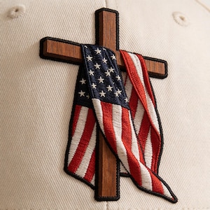 May include: A beige baseball cap featuring an embroidered wooden cross with an American flag draped over it. The flag has red and white stripes and blue field with white stars. The cross is made of dark wood and has a black border.