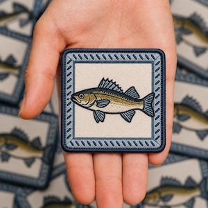 May include: A square embroidered patch featuring a central illustration of a fish with olive green and dark blue details. The fish is set against a light beige background and framed by a dark blue border with a diagonal stitched pattern.