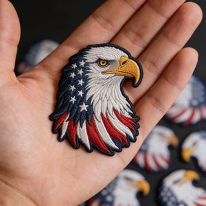 May include: Embroidered patch featuring an American bald eagle head. The patch showcases the eagle's head with white, blue, and red colors, representing the American flag. The eagle has a yellow beak and eye.