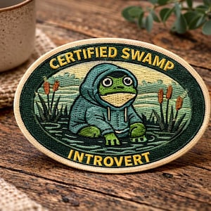May include: An oval patch with the text "CERTIFIED SWAMP INTROVERT" in gold lettering. The patch features a cartoon frog wearing a blue hoodie, sitting in a swamp. The patch is surrounded by a green border and is on a wooden surface.