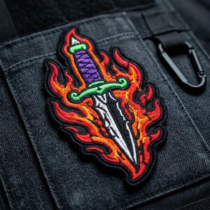 May include: An embroidered patch featuring a silver dagger with a purple handle and a bright green crossguard. The dagger is surrounded by stylized flames in shades of red, orange, and yellow, all outlined with a thick black border.