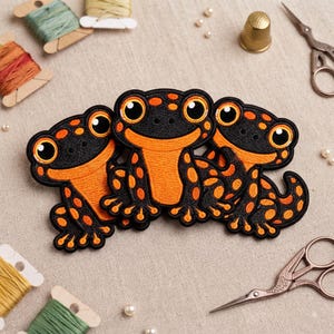 May include: Three embroidered patches featuring stylized frogs with black and orange coloring. Each frog has large, round eyes, a wide smile, and spotted patterns on the body. The patches are arranged in a cluster with black stitched borders.