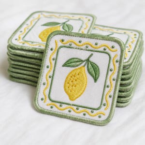 May include: A stack of square embroidered patches featuring a yellow lemon with two green leaves in the center. The design is framed by a wavy yellow line and a green border with small green dots on a white background with stitched edges.