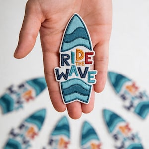 May include: Embroidered surfboard patch with the words "Ride the Wave" in blue, red, and gold. The surfboard design features blue waves. Additional patches are visible in the background.
