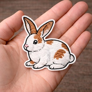 May include: Embroidered patch of a white and brown rabbit with a black outline. The rabbit has brown ears and spots on its back. The patch is held in a hand.