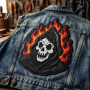 May include: A blue denim vest featuring a large embroidered patch on the back. The patch depicts a white skull wearing a black hooded cloak, surrounded by vibrant orange and red flames with a thick black border stitched onto the fabric.