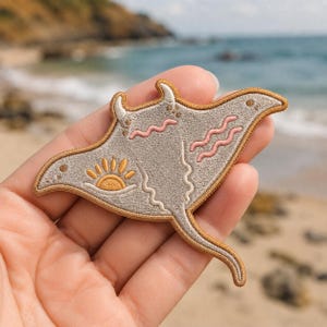 May include: Embroidered patch featuring a stylized manta ray design. The patch has a light gray body with a gold border and accents. It includes a sun graphic and pink wave details, perfect for adding to clothing or accessories.