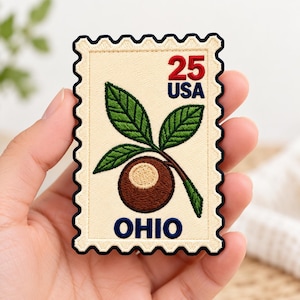 May include: A rectangular embroidered patch shaped like a postage stamp with a cream background and a dark blue scalloped border. It features a brown buckeye nut with green leaves, the red number 25, blue text reading USA, and blue text reading OHIO.