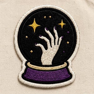 May include: An embroidered patch shaped like a crystal ball with a black background, featuring a white hand with purple fingernails reaching upward. The design includes yellow stars, small dots, and a purple base with a gold rim, all outlined in white.