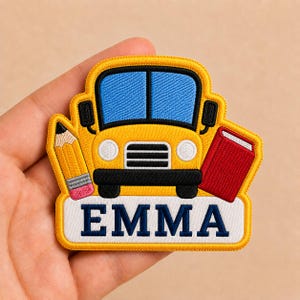 May include: Embroidered patch featuring a yellow school bus with blue windows, a red book, and a pencil. The patch has the name "EMMA" in navy blue letters on a white background. The patch is outlined in yellow.