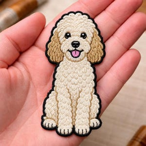 May include: An embroidered patch shaped like a sitting dog with curly cream-colored fur, tan ears, black eyes, a black nose, and an open mouth showing a pink tongue. The entire design is outlined in a thick black border with detailed stitching.