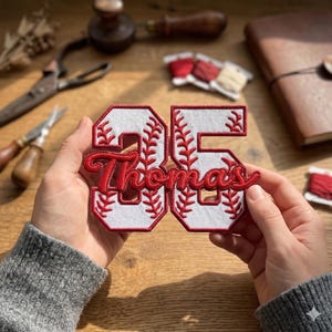 May include: A baseball-themed patch with the number "35" in a red and white design. The name "Thomas" is embroidered in red script across the numbers. The patch is held in front of a wooden surface.