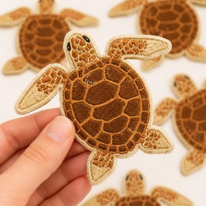 May include: Embroidered sea turtle patches in shades of brown and tan. The patches feature detailed stitching, with a textured shell design and outstretched flippers. The patches are likely for decorative purposes, such as on clothing or bags.