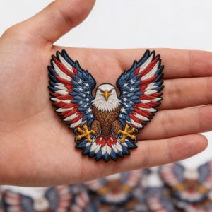 May include: Embroidered patch featuring a bald eagle with outstretched wings. The eagle's wings display the American flag design with red and white stripes and blue stars. The eagle's body is brown, with a white head and yellow beak and talons.
