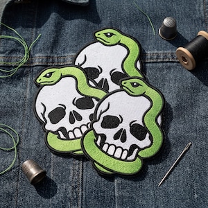 May include: Three embroidered patches featuring white skulls with black eye sockets and teeth, each wrapped by a bright green snake with a black outline. The patches are arranged in a cluster on a dark blue denim fabric background with sewing tools.