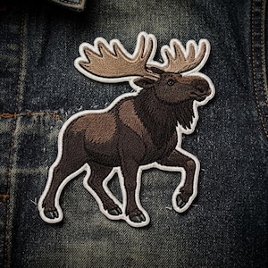 May include: An embroidered patch featuring a brown moose with large antlers, depicted in profile. The moose has dark brown fur on its body and lighter brown fur on its antlers, outlined with a white border against a dark blue denim fabric.