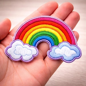 May include: Embroidered rainbow patch featuring a vibrant spectrum of colors: red, orange, yellow, green, blue, and purple. The rainbow is arched over two fluffy white clouds with purple and blue outlines. This patch is ideal for adding flair to clothing or accessories.