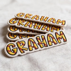 May include: A stack of embroidered patches with the name "GRAHAM" in yellow letters with red stitching, resembling a baseball. The patches have a cream-colored background and a black border.