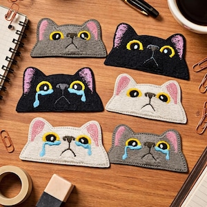 May include: Embroidered cat face patches in gray, black, and white, with pink ears and yellow eyes. Some patches feature blue tear drops. The patches are arranged on a wooden surface, with office supplies in the background.