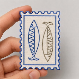 May include: A rectangular embroidered patch shaped like a postage stamp with a scalloped blue border. The off-white fabric features two stylized fish designs: one outlined in blue with a chevron pattern and one in tan with a scale pattern.