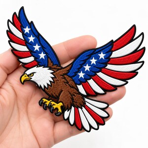 May include: An embroidered patch featuring a bald eagle in flight. The eagle has a brown body, a white head, and a yellow beak and talons. Its wings are patterned with red and white stripes and a blue section adorned with five white stars.