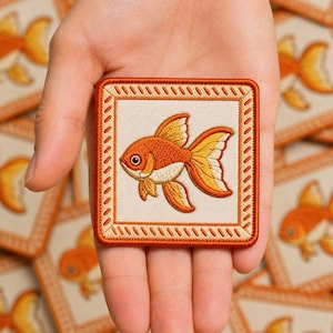 May include: A square embroidered patch featuring a goldfish with orange and yellow scales on a cream background. The patch is framed by a decorative border with a repeating diagonal stitch pattern in shades of orange and light brown.