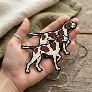 May include: Two embroidered patches shaped like dogs with white fur and brown spots. The patches feature detailed stitching with black outlines, depicting two dogs standing in profile, one slightly behind the other, showcasing a canine design.