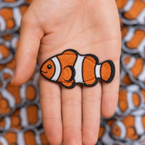 May include: An embroidered patch shaped like a clownfish with vibrant orange and white stripes, outlined in black thread. The fish features a small black eye and a simple stitched smile, showcasing detailed needlework throughout the design.