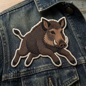 May include: An embroidered patch featuring a brown wild boar in a running pose with dark brown fur, pointed ears, and small tusks. The patch has a cream-colored border and is attached to a dark blue denim jacket with visible stitching and a metal button.