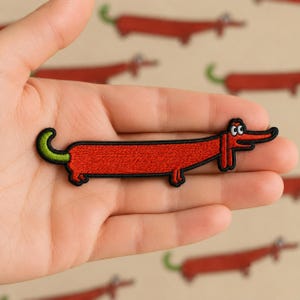 May include: A red embroidered dachshund patch with a black outline and a green tail. The cartoon dog has a long body and is held in a hand. The background features a pattern of similar dachshunds.