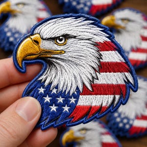 May include: An embroidered patch featuring the profile of a bald eagle head. The eagle has white feathers, a yellow beak, and an eye with a dark pupil. The lower portion of the patch displays a blue field with white stars and red and white stripes.