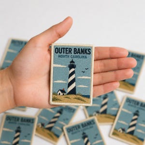 May include: A rectangular embroidered patch featuring a blue sky, a black and white striped lighthouse, and a small house on a sandy dune. The text reads OUTER BANKS NORTH CAROLINA in dark blue letters. The patch has a cream-colored border.