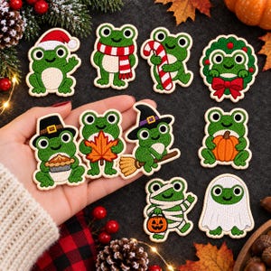 May include: A collection of ten embroidered iron-on patches featuring green frogs in various seasonal costumes, including a Santa hat, a red and white scarf, a candy cane, a holiday wreath, a pilgrim hat with pie, an autumn leaf, a witch hat, a pumpkin, a mummy, and a ghost.